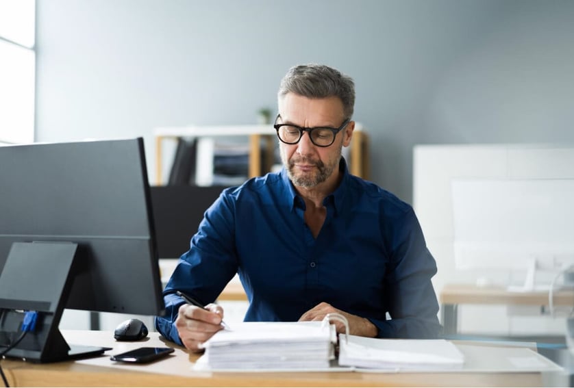 Office worker in a blue shirt sat at a desk looking through a folder of paperwork